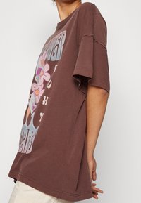 Camiseta oversized marrón de algodón, con gráficos florales coloridos y texto. Mangas cortas, cuello redondo y corte relajado.