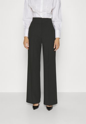 Kobieta ubrana w czarne szerokie spodnie z wysokim stanem, białą koszulę zapinaną na guziki oraz czarne szpilki z wydłużonym noskiem, stojąca na białym tle.