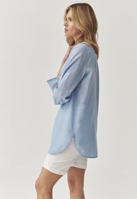 Camicia a maniche lunghe azzurro chiaro in morbido tessuto con orlo curvo. Indossata sopra pantaloni corti bianchi, mostra un fit rilassato e un design casual.