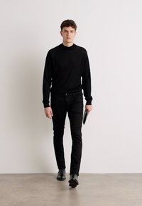Pull en maille noir, jean slim noir et chaussures en cuir noir. Modèle tenant une pochette, se tenant devant un fond uni.
