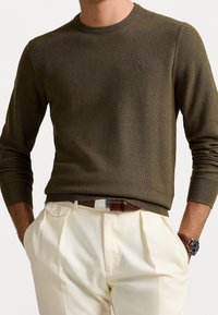 Olivgrön stickad tröja med rund halsringning, ribbade ärmslut och fåll. Tillsammans med beige byxor och ett brunt bälte med en silverspänne.