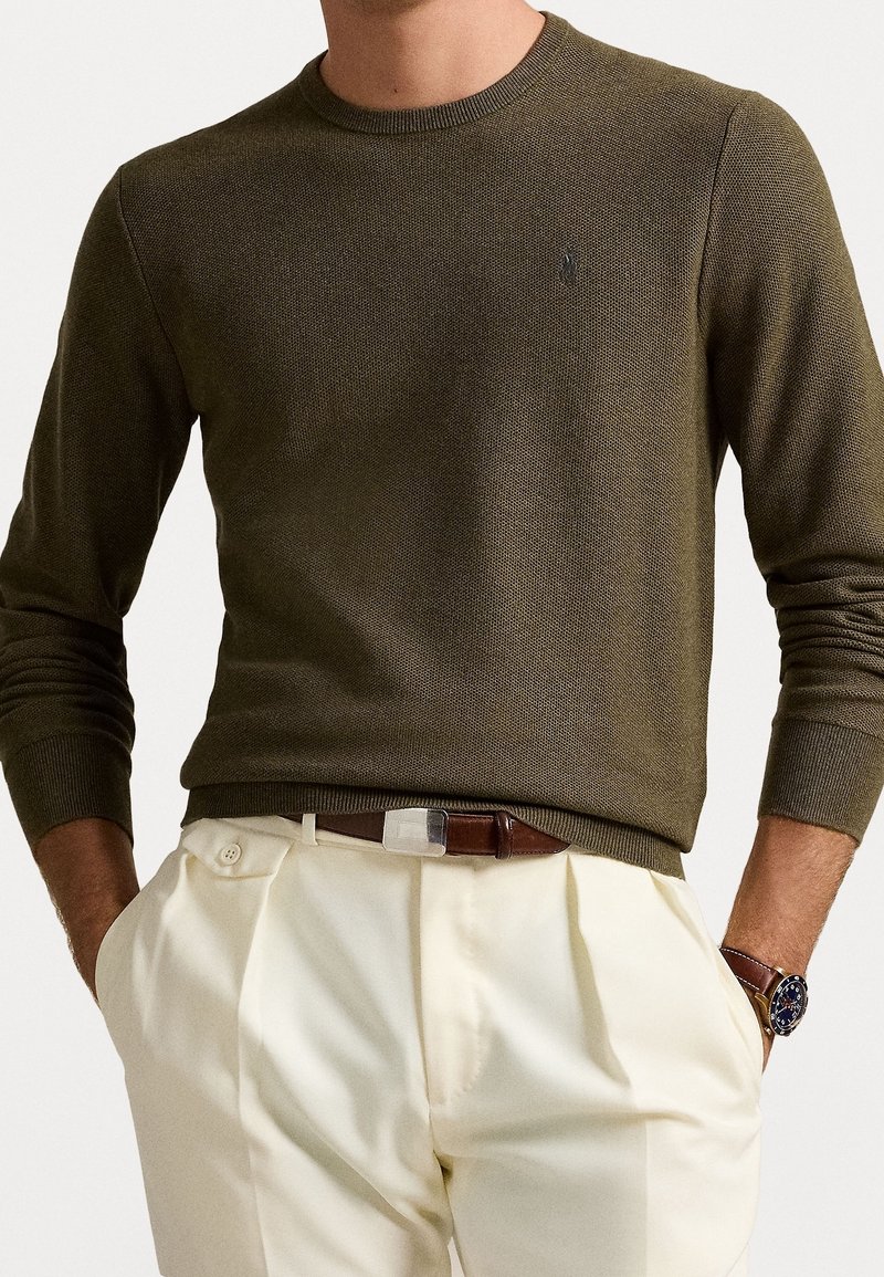 Olivgrön stickad tröja med rund halsringning, ribbade ärmslut och fåll. Tillsammans med beige byxor och ett brunt bälte med en silverspänne.