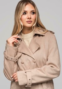 Beżowy płaszcz dwurzędowy wykonany z miękkiego materiału przypominającego zamsz, z pagonami, szerokimi klapami i ozdobnymi guzikami; paski na rękawach z metalowymi elementami.