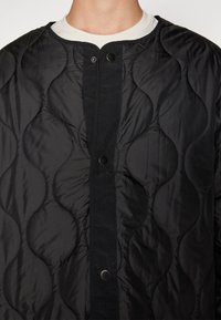 Quilted black jacket with a button closure, featuring a textured pattern, round neckline, and smooth fabric for a sleek appearance.