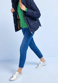 Marineblaues, gesteppte Jacke, grüner Cardigan, Jeans und silberne Sneakers. Fokus auf Texturen und Farbkontraste. Lässiges Outfit mit klaren Schichten.