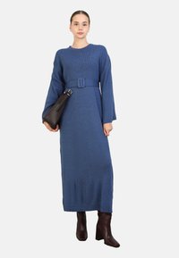 Blaues geripptes langes Kleid mit einem Gürtel, mit langen Ärmeln und gerader Silhouette. Kombiniert mit braunen Absatzstiefeln und einer dunklen Handtasche.