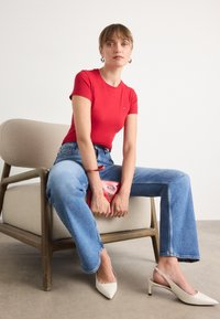 T-shirt rouge ajusté, jeans bleu droit, escarpins slingback blancs à bout pointu, tenant un sac rouge. Design minimaliste, textures variées.