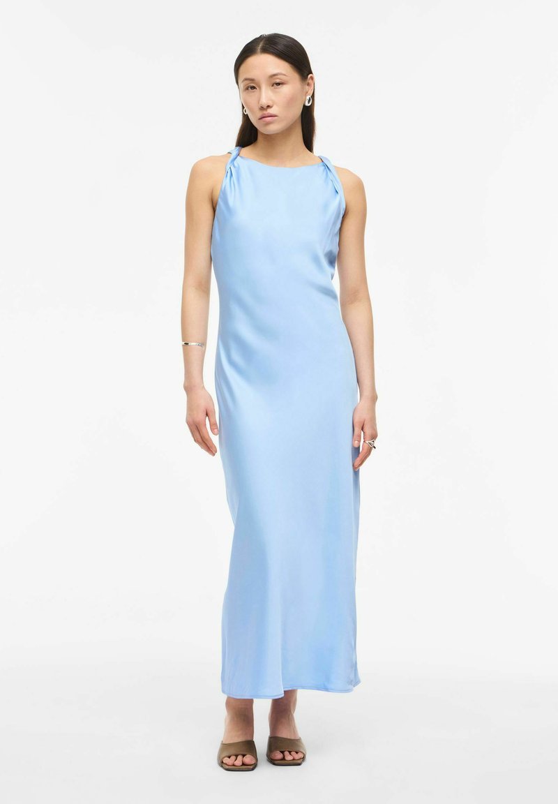 Femme portant une robe maxi en satin bleu clair sans manches avec des liens aux épaules, des boucles d'oreilles créoles argentées et des talons ouverts taupe, debout sur fond blanc.