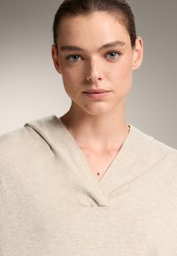 Beige gebreide trui met een V-hals en een capuchon. Lichtgewicht textuur, met een schoon ontwerp en subtiele stiksels.