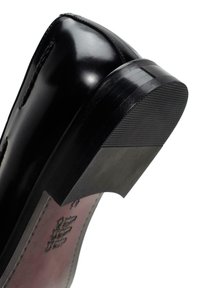 Chaussure en cuir noir avec une finition brillante, dotée d'un bout arrondi et d'une semelle en caoutchouc texturée. Des coutures de haute qualité sont visibles.