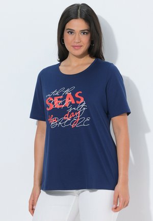 Junge Frau mit dunklem Haar, die ein dunkelblaues T-Shirt mit rotem und weißem Schriftzug trägt, kombiniert mit weißer Hose, stehend vor weißem Hintergrund.