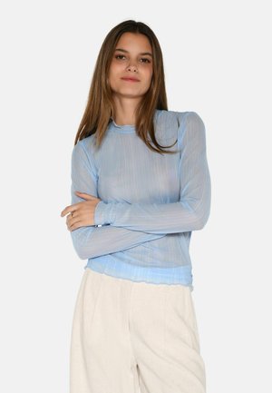 Top lungo a maniche lunghe di un blu chiaro trasparente con strisce orizzontali testurizzate, scollatura arricciata e orlo scalloppato. Indossato con pantaloni beige.