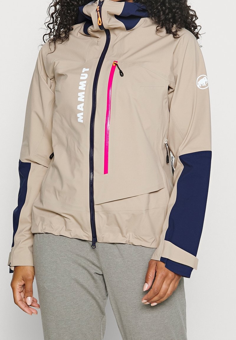 Chaqueta de poliéster beige con detalles en azul marino, presenta un collar alto, bolsillos con cremallera y una llamativa cremallera rosa. Logo bordado en la manga.