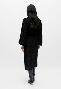 Kvinde med langt mørkt hår iført en sort midikjole med blomstertekstur og sorte knæhøje støvler med hæl, stående mod en ensfarvet hvid baggrund.