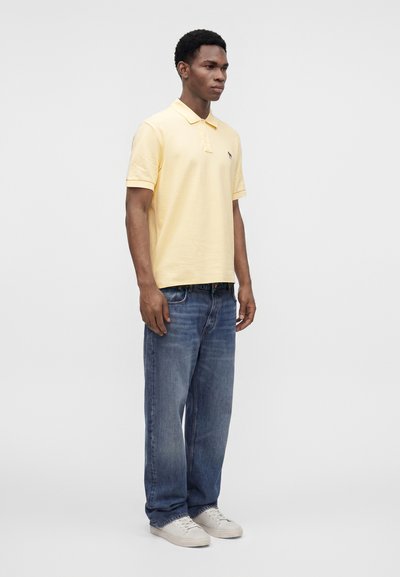 Homme portant un polo jaune pâle, un jean bleu ample et des baskets blanches, debout devant un fond uni gris clair.