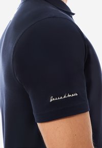 Marineblaue Polohemden aus strukturiertem Stoff mit kurzen Ärmeln. Der linke Ärmel ist mit weißem, gesticktem Text "Gassadi'mare" versehen.
