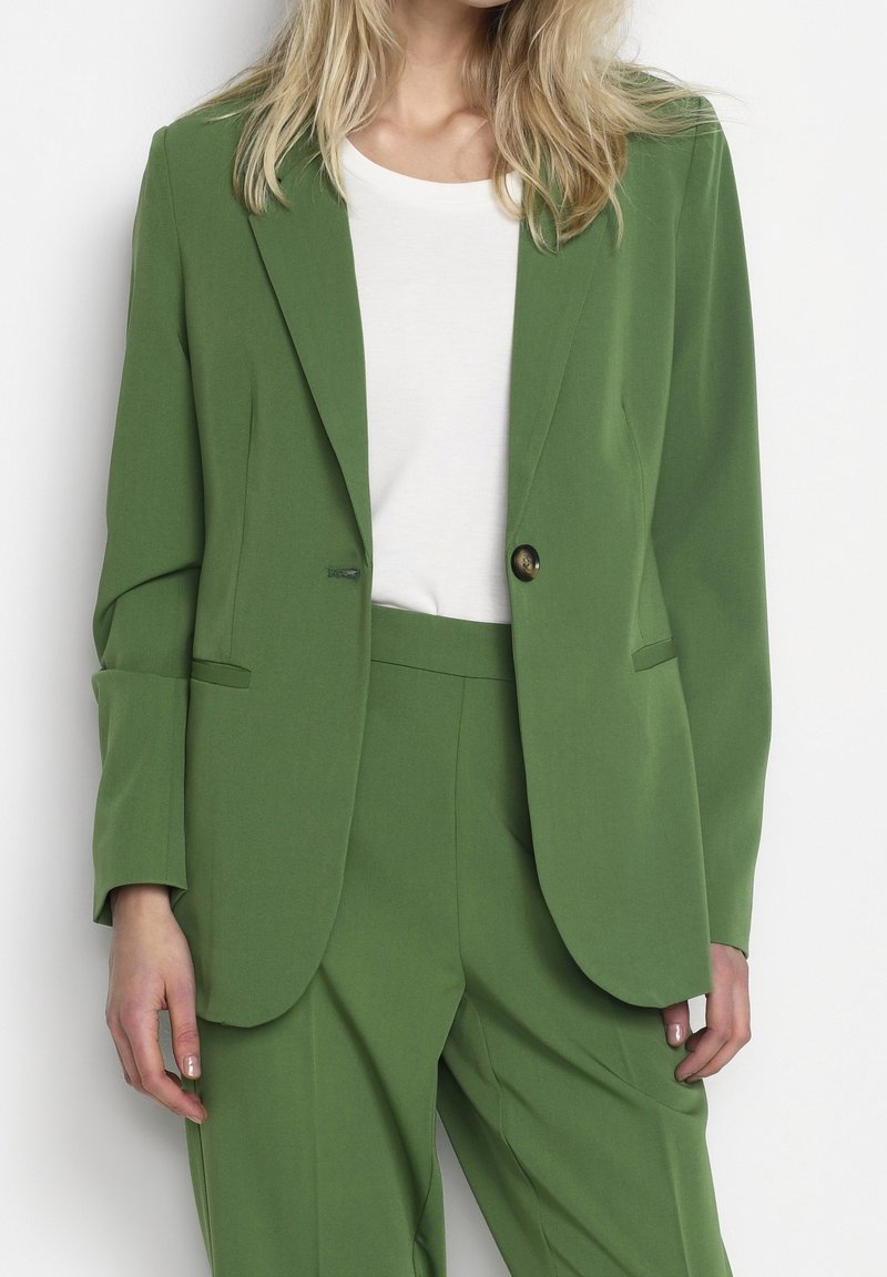 Femme blonde portant un costume vert avec un blazer à un bouton et un pantalon assorti sur un t-shirt blanc à col rond.