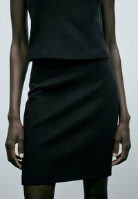 Robe noire cintrée avec une finition texturée. Haut court sans manches associé à une jupe longueur genou. Lignes simples et épurées partout.