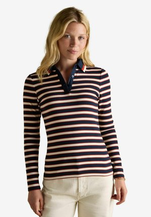 Blonde woman wearing a navy, white, and red striped long-sleeve polo shirt with cream pants, standing against a plain background.