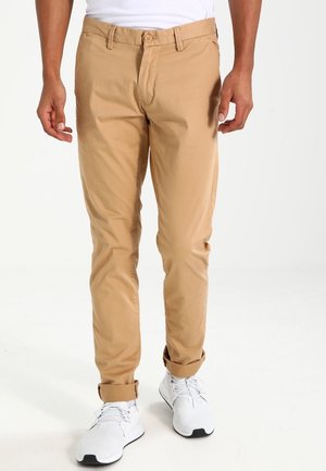 Mężczyzna w beżowych, dopasowanych spodniach podwiniętych u dołu i białych trampkach, stojący na tle jednolitej białej ściany.