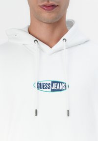 Bijela dukserica s kapuljačom s prednjim izvezenim logotipom "GUESS JEANS" u plavoj i zelenoj boji. Ima vezice s metalnim srebrnim vrhovima.