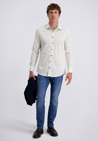 Camicia bianca a maniche lunghe con pois neri, colletto a camicia, abbinata a jeans blu e scarpe scure, mentre tiene in mano un blazer blu navy.