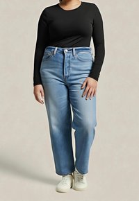 Personne portant un jean taille haute bleu clair, un t-shirt noir à manches longues et des baskets blanches, debout devant un fond clair uni.