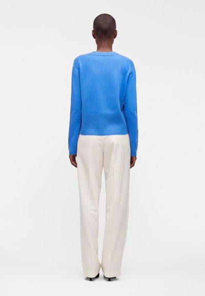 Personne debout de dos, portant un pull bleu vif et un pantalon blanc ample sur un fond clair et uni.
