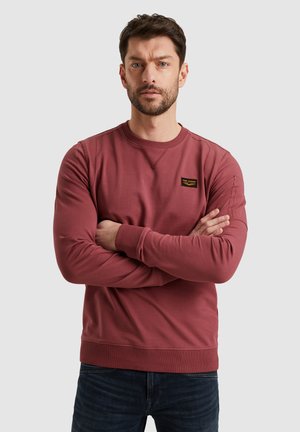 Felpa bordeaux realizzata in morbido tessuto, con maniche lunghe, polsini e orlo a coste; presenta una piccola patch nera con logo sul petto.