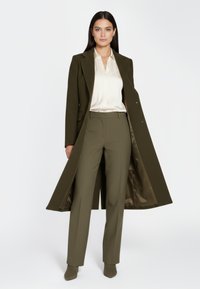 Olijfgroene op maat gemaakte jas over een crèmekleurige blouse, met bijpassende olijfgroene broek en suède enkellaarzen, met een strak en verfijnd ontwerp.