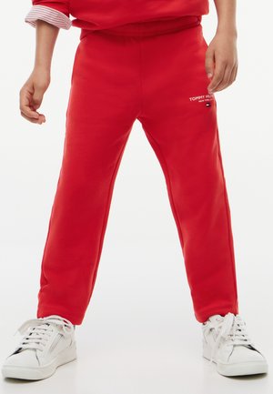Barn iført røde Tommy Hilfiger joggingbukser og hvide sneakers står med benene fra hinanden på en lys baggrund.