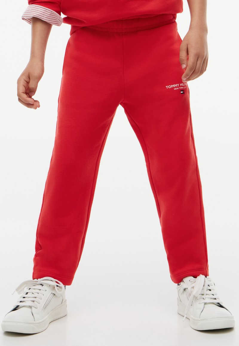 Παιδί με κόκκινη φόρμα Tommy Hilfiger και λευκά αθλητικά παπούτσια στέκεται με τα πόδια ανοιχτά σε φωτεινό φόντο.