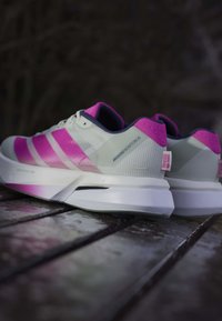 Hvide og lyserøde Adidas løbesko på en træoverflade, der viser siden og bagsiden med synlig branding og polstrede såler.