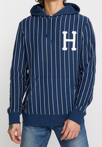 Mörkblå randig hoodie med vita vertikala ränder, framficka och en stor vit "H"-logotyp på bröstet. Bomullsblandad tyg.