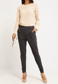 Beige satijnen blouse met lange mouwen en knoopdetails, gecombineerd met donkergrijze tapered broek en nude hoge hakken. Houdt een witte clutch tas vast.