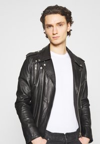 Veste en cuir noir de motard avec poches zippées et détails d'épaulettes, portée sur un t-shirt blanc uni, sur un fond neutre.