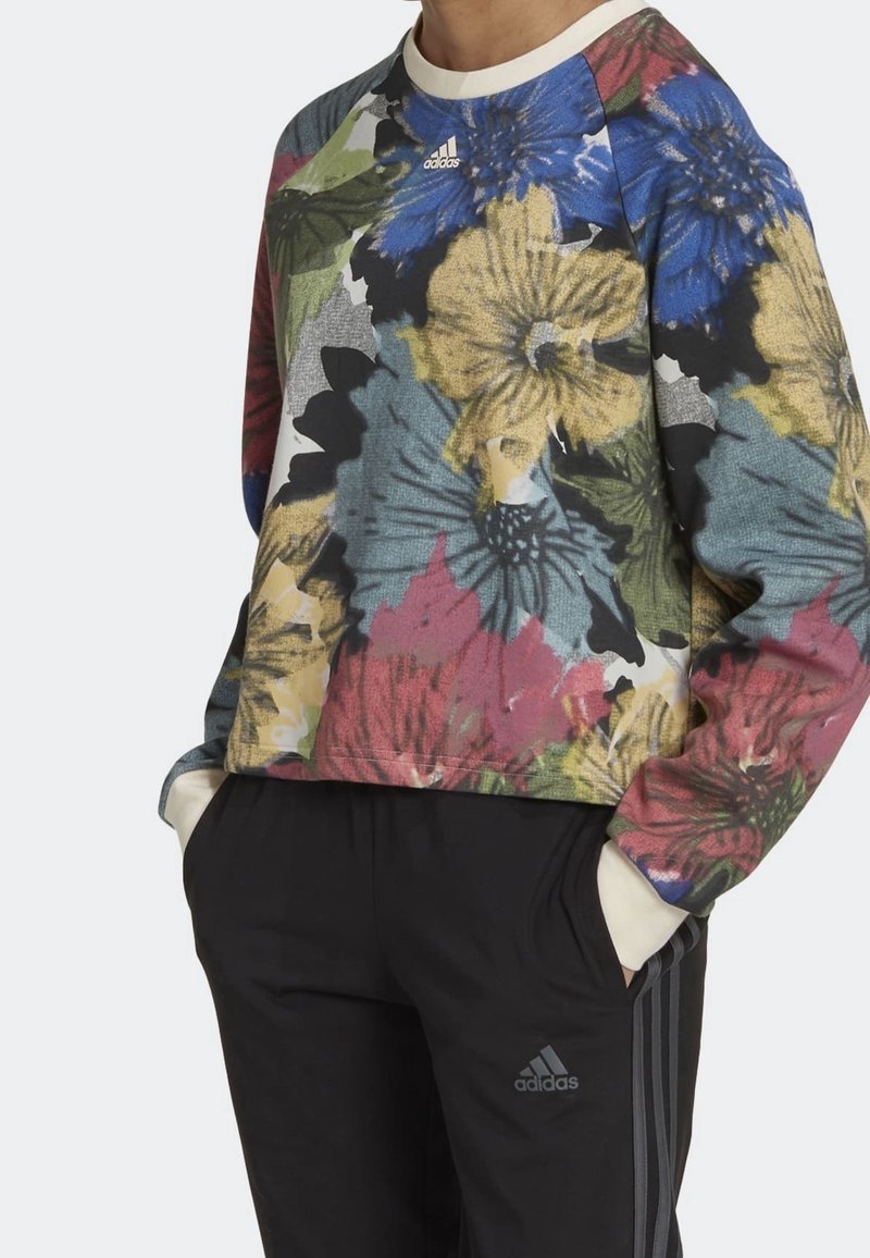 Personne portant un sweat Adidas à manches longues à fleurs multicolores et un pantalon de survêtement Adidas noir, les mains dans les poches.