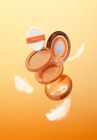 Kompaktowy produkt kosmetyczny w pomarańczowym opakowaniu, z aplikatorem gąbkowym, wypełnionym kolorowym podkładem oraz lustrzanym wnętrzem pokrywki z logo marki.