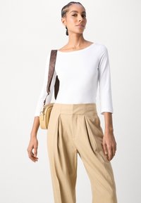 Top blanco de manga larga combinado con pantalones de taza beige con pliegues. El modelo lleva un pequeño bolso marrón con una correa de patrón marrón.
