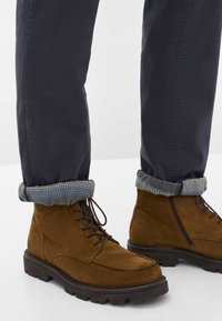 Stivaletti in suede marrone con finitura testurizzata, punta tonda e suole in gomma nera. Indossati con pantaloni blu navy con orli a scacchi.