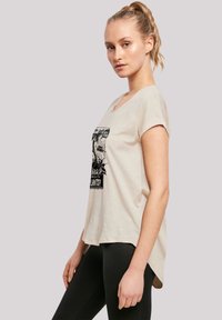 Beige short-sleeve T-shirt made of soft fabric, featuring a black graphic print at the center. Curved hemline, paired with black leggings.
