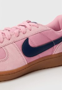Roza tenevka iz semiša in platna, z temno modrim Nike swooshom in rjavo gumijasto podplatom. Zavezovanje z vezalkami in teksturirani dodatki.