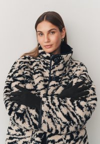 Műszőrme kabát fekete és bézs tigriscsík mintával, magas nyakkal és fekete gombokkal. A modell fekete kesztyűt visel. Semleges háttér.