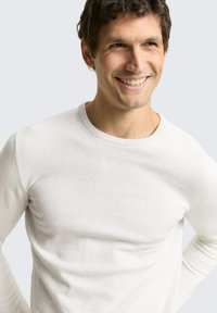 Leichter weißer Pullover mit Rundhalsausschnitt, geripptem Kragen, glatter Textur und figurbetontem Schnitt, getragen von einem lächelnden männlichen Modell vor einem schlichten Hintergrund.