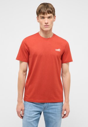 Young man standing facing forward wearing a plain orange short-sleeve t-shirt with a small white horse logo and blue jeans.