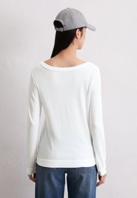 Camicia bianca a maniche lunghe con scollatura ampia e bordo a costine, abbinata a jeans in denim scuri. Il modello indossa un cappellino da baseball grigio.