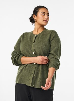 Woman with dark hair in a bun wearing gold hoop earrings and an olive green buttoned cardigan, adjusting one sleeve against a white background.