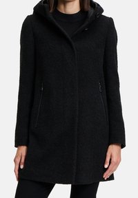 Cappotto nero in misto lana con finitura testurizzata, cappuccio e tasche laterali con zip. Presenta un taglio dritto e chiusura con bottoni.