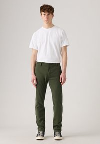 Young man standing facing forward wearing a plain white t-shirt, olive green pants, and black high-top sneakers.
