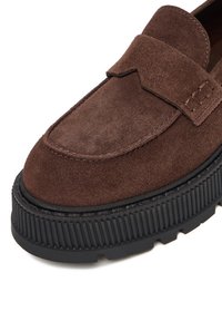 Loafers din suede maro cu o talpă groasă din cauciuc negru, având un vârf cusut și un detaliu pliat mic la vamp pentru un plus de textură.